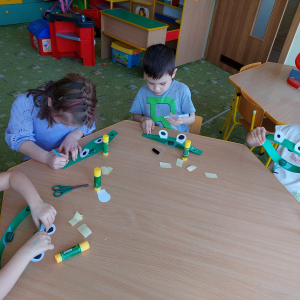 Dzieci siedzą przy stoliku i przyklejają elementy. 