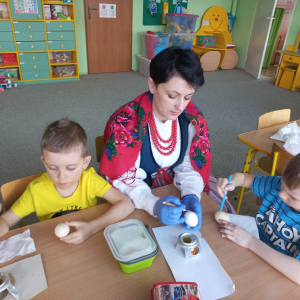 Dzieci siedzą przy stolikach i ozdabiają jajka metodą batikową.