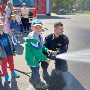 Chłopiec wspólnie ze strażakiem trzyma wąż strażacki i polewają wodą