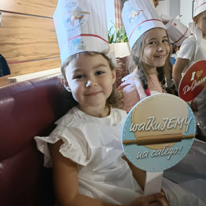 Dziewczynki siedzą ubrane w fartuszki i czapki kucharskie