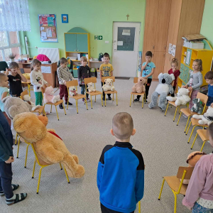 Dzieci stoją w kole a przed nimi na krzesełkach siedzą misie