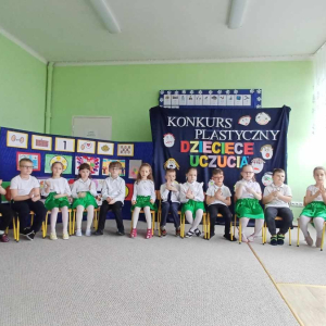 Dzieci siedzą na krzesełkach i pokazują gesty do piosenki