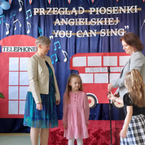 Dziewczynka z Panią Dyrektor stoi przed sceną i wręczana jest jej nagroda oraz dyplom. 