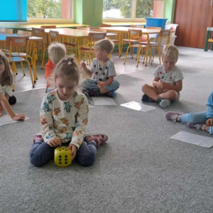 Dzieci siedzą na dywanie a w środku koła dziewczynka trzyma kostkę