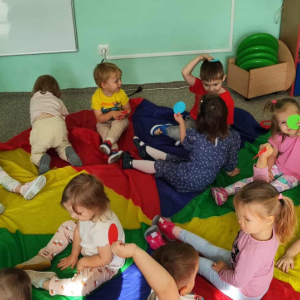 Dzieci siedzą na chuście animacyjnej i trzymają w rękach kolorowe kropki
