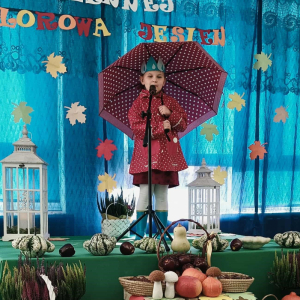 Dziewczynka stoi na scenie, trzyma parasol i śpiewa piosenkę.