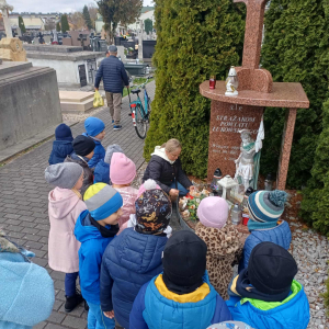 Dzieci stoją przed grobem strażaków a pani zapala znicz.