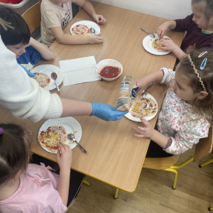 Dzieci siedzą przy stoliku i przygotowują swoje pizze.