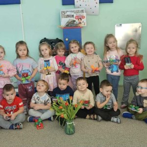 Dzieci z grupy Biedroneczki pozuja do zdjęcia, dziewczynki stoją i trzymają kwiatkia chłopcy siedzą