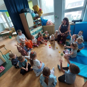 Dzieci siedzą w kole, na podłodze leżą kolorowe piłeczki. 