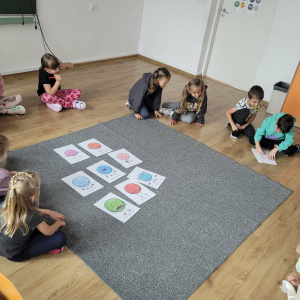 Dzieci siedzą w kole. Na dywanie leżą kolorowe obrazki z kropkami. 