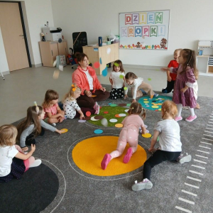 Dzieci siedzą na dywanie z panią, na dywanie leżą kolorowe kropki.