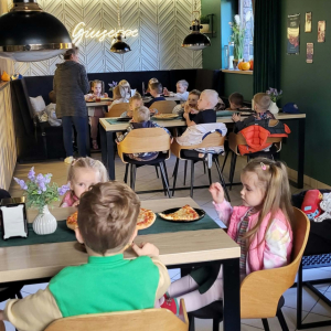 Dzieci siedzą przy stolikach i jedzą pizzę.