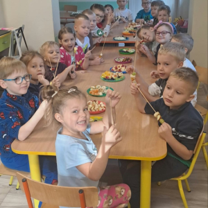Dzieci siedzą przy stoliku i robią szaszłyki z owoców